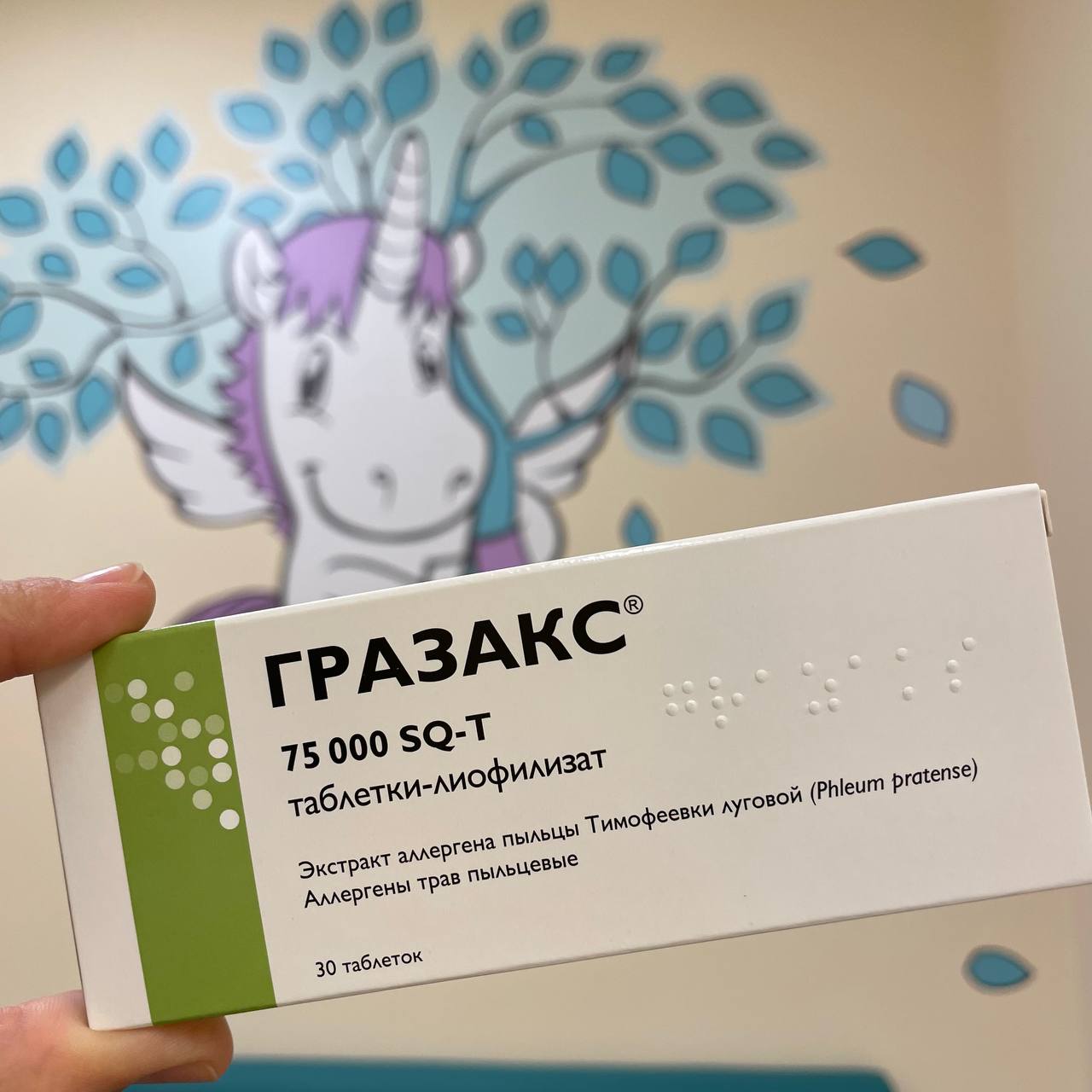 Упаковка препарата для АСИТ «Гразакс» (таблетки-лиофилизат с экстрактом пыльцы тимофеевки луговой) в руке на фоне настенного рисунка с единорогом в кабинете